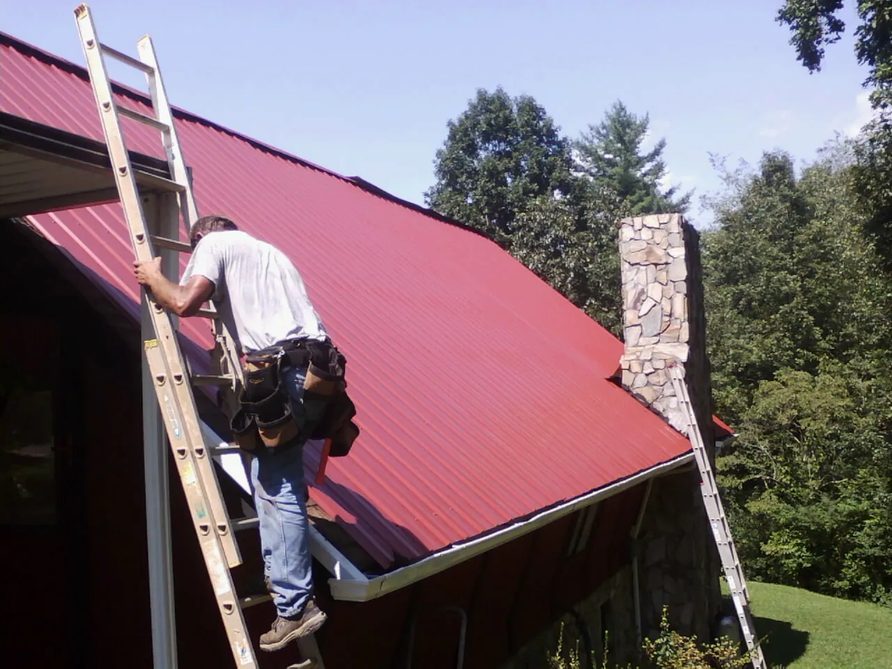 Roofer on a ladder beside a red metal roof and stone chimney — Soffit & Fascia Repair by Roofing Brothers in Capon Bridge